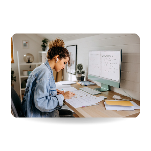 There is a girl in a desk studying in front of the computer and writing in a notebook